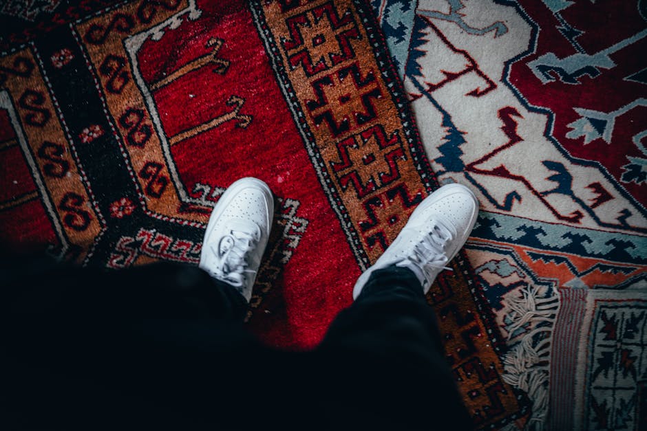 A person wearing casual clothing is kneeling on a patterned carpet in a room, placing a white cloth into a yellow and black vacuum cleaner with a flexible hose. The flooring is a decorative, patterned