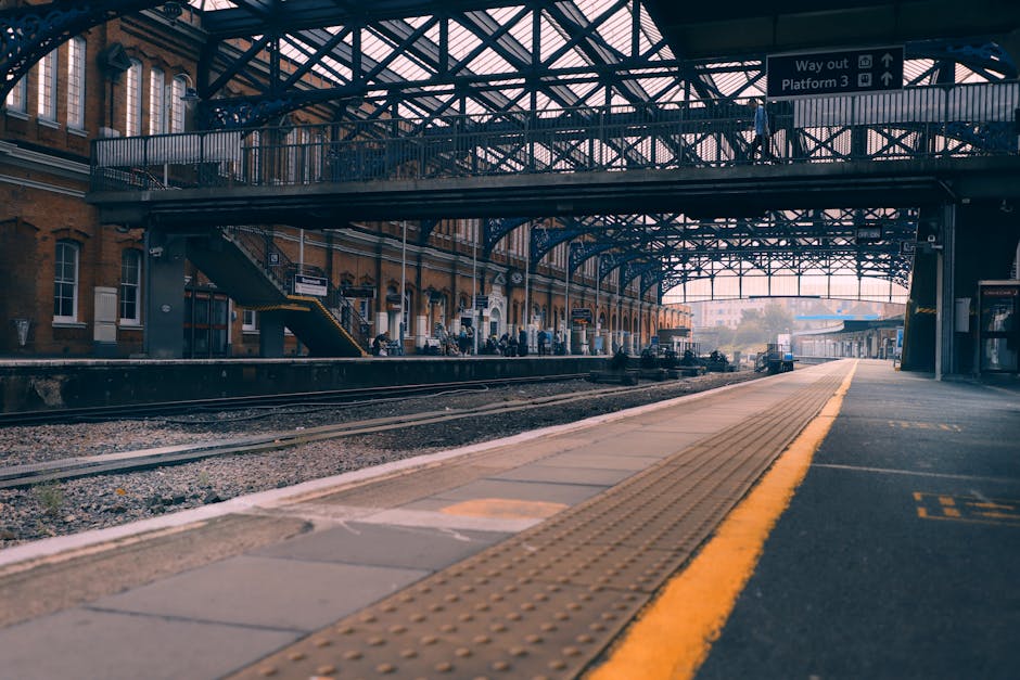 View of a train station platform with a yellow tactile strip along the edge, adjacent to railway tracks. The platform surface is clean and smooth, made of concrete and ceramic tiles, with some areas a