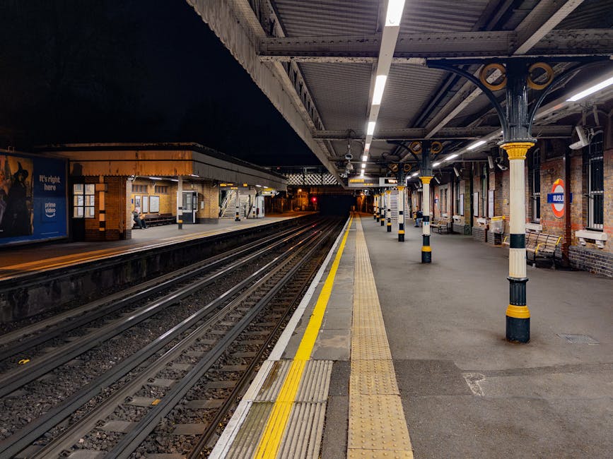 View of a train station platform with a yellow tactile strip along the edge, adjacent to railway tracks. The platform surface is clean and smooth, made of concrete and ceramic tiles, with some areas a